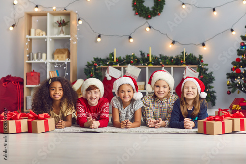 Group of happy kids in Santa hats looking at camera lying on floor in cozy living-room with presents