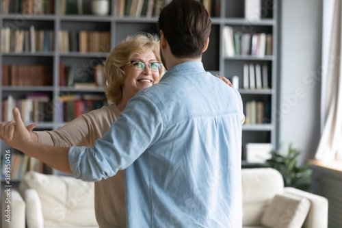 Back rear view affectionate caring young man leading in slow dance smiling middle aged 60s senior mother at home. Happy proud loving old mature woman enjoying waltz with grownup son in living room.