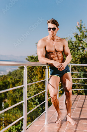 Fashion Portrait Of A Muscular Man posing shirtless in swim trunks and sunglasses. Rich Muscular handsome man relax on his villa outdoors