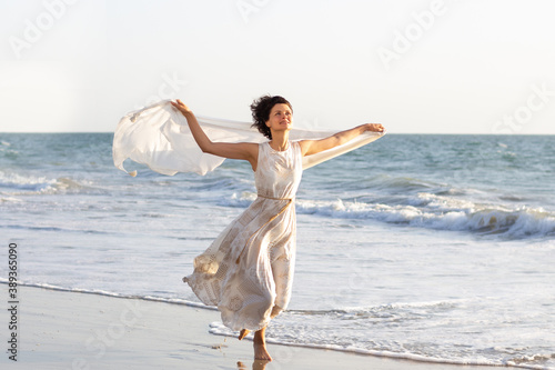 a beautiful girl in a white dress walks along the seashore with a pareo in her hands