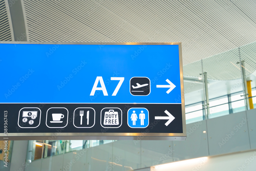 Shield with signs indicating airport amenities and public utilities, and arrow pointing to gate to board the plane for departure hangs on wall, close up. Shield with signs indicating airport amenities and public utilities, and arrow pointing to gate to board the plane for departure hangs on wall, close up.