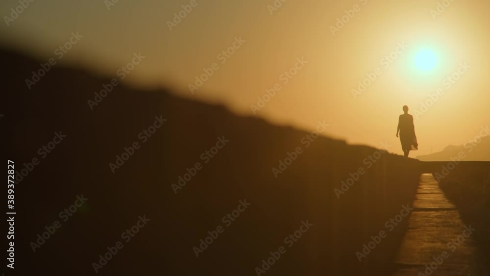 Woman in a red dress and a straw hat walks along the edge front of a cliff by the sea alone at sunset Woman in a red dress and a straw hat walks along the edge front of a cliff by the sea alone at sunset