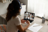 Rear view businesswoman wearing headphones working from home, engaged in online conference, sitting at work desk with laptop, diverse colleagues brainstorming by group video call, briefing