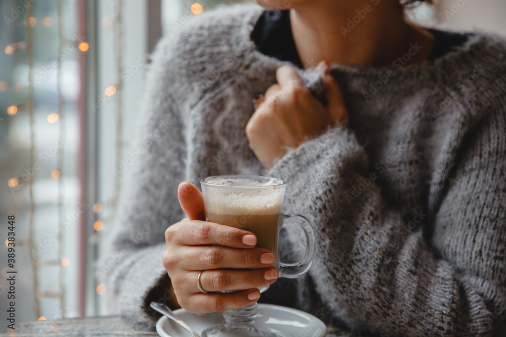 A young brown-haired woman in a warm jacket is sitting in a cafe at a table by the window and holding a glass of coffee A young brown-haired woman in a warm jacket is sitting in a cafe at a table by the window and holding a glass of coffee