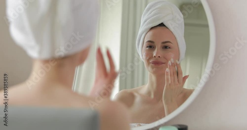 Good-looking young woman with white towel on head applying cream on her face after shower. Charming female doing morning procedures for skin care.