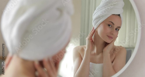 Attractive young female with bare shoulders viewing her face skin while standing at bathroom. Reflection in mirror. Concept of skin care.