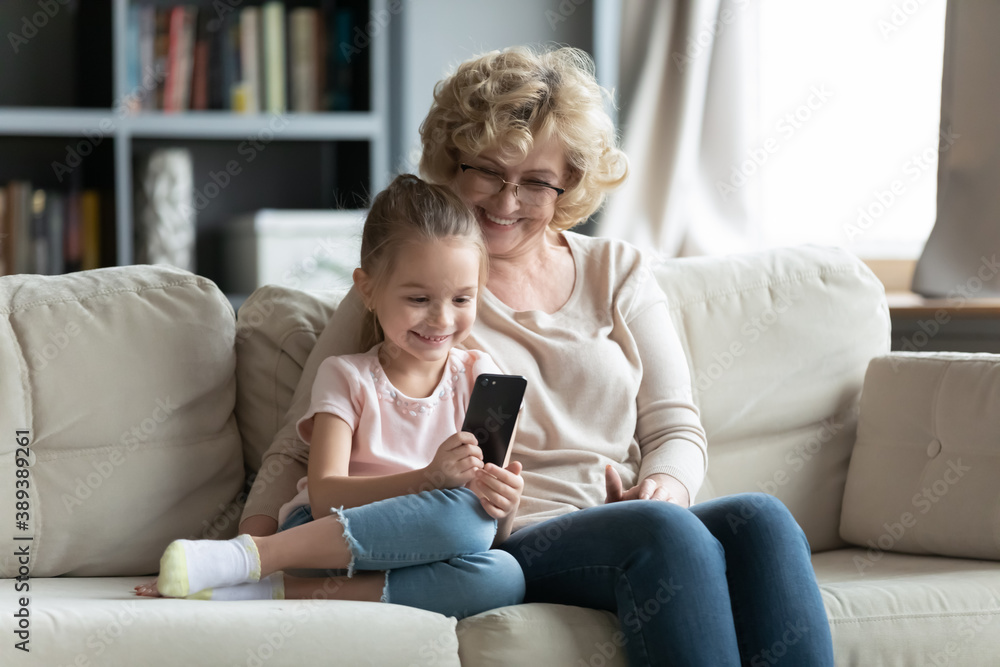 Happy small 6s cute kid girl using smartphone applications, sitting on sofa with caring elderly senior grandmother in eyeglasses. Smiling different female generations family playing mobile games. Happy small 6s cute kid girl using smartphone applications, sitting on sofa with caring elderly senior grandmother in eyeglasses. Smiling different female generations family playing mobile games.