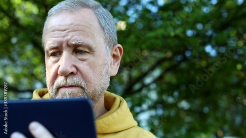 Senior bearded man using tablet for communication looking at screen resting outdoor enjoying autumn spring sunny nature. Sunny background. Internet connect. Free communicating with family.