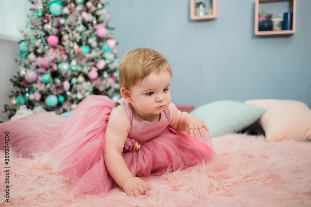 a playful little girl in a festive pink dress crawls on a furry pink blanket on the background of a Christmas tree a playful little girl in a festive pink dress crawls on a furry pink blanket on the background of a Christmas tree