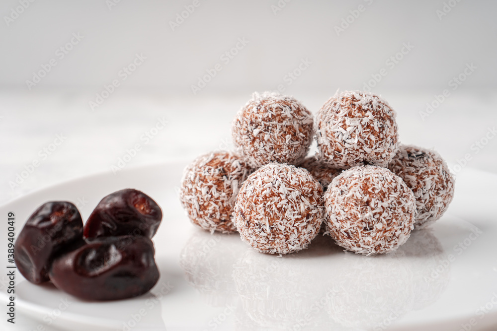 Candy balls made of dates and coconut shavings in a white dish on a light background near the dates Candy balls made of dates and coconut shavings in a white dish on a light background near the dates