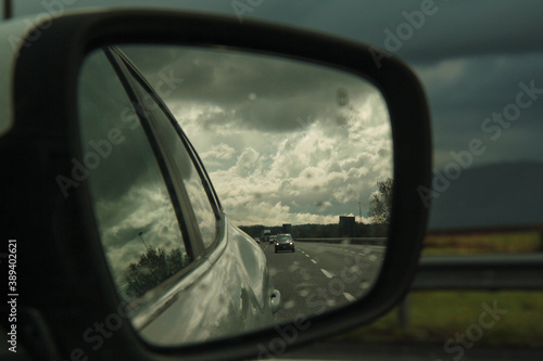 view of the road, reflected from the side mirror of the car, under a blue and cloudy sky © Valentina