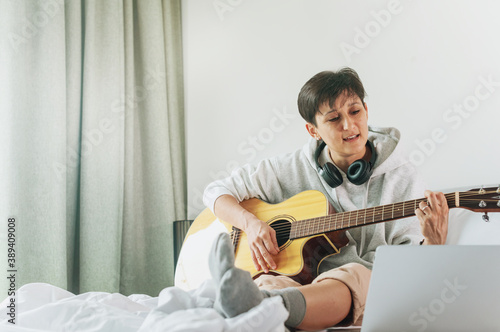 Lady with is singing song and playing on acoustic guitar with laptop on bed in apartment.