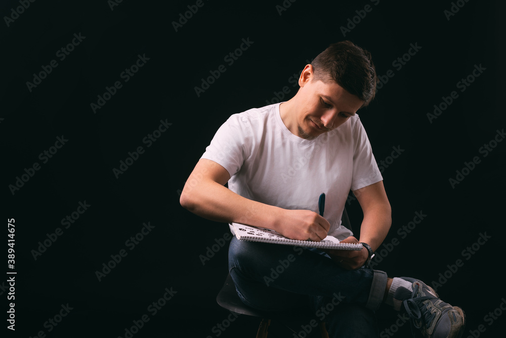 A picture of a man drawing on a paper sitting on a chair near a black wall . A picture of a man drawing on a paper sitting on a chair near a black wall .