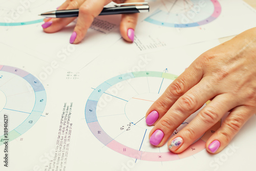 Workplace of a modern astrologer. Printouts of astrological charts, charts, tables, diagrams lying on the table. Hands of a woman astrologer.