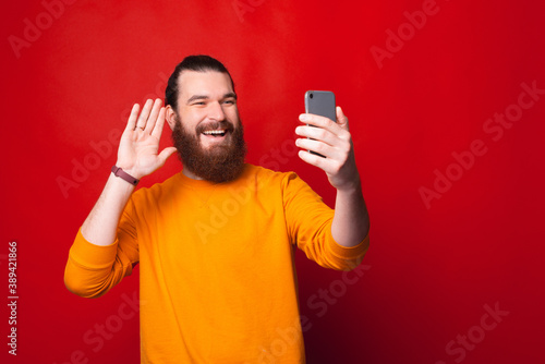 A bearded young man is smiling and waving at his phone having a video call . © Vulp