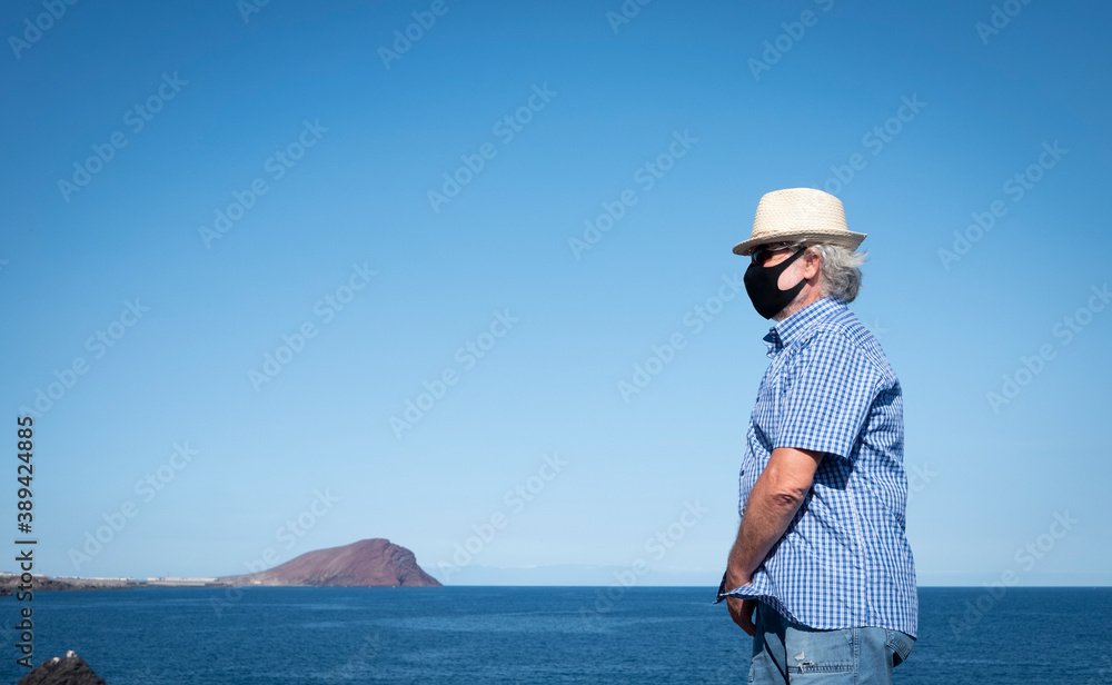 Old man white haired standing on the cliff enjoying vacation wearing face mask because of Covid-19 coronavirus. Blue sea, mountain and island Gran Canaria behind him