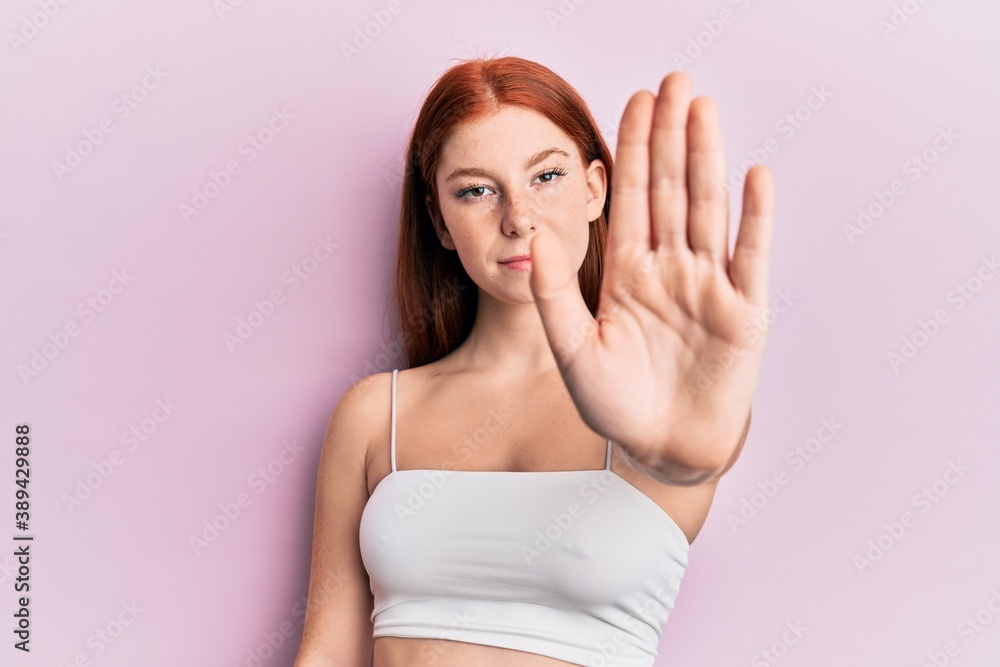 Young red head girl standing over pink background with open hand doing stop sign with serious and confident expression, defense gesture Young red head girl standing over pink background with open hand doing stop sign with serious and confident expression, defense gesture