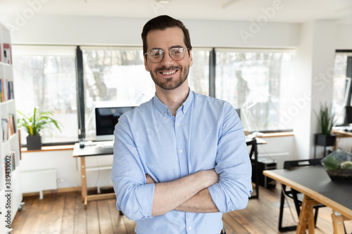 Portrait of happy handsome 30s businessman in eyeglasses standing in modern creative workplace with folded hands, successful startup male entrepreneur owner showing confidence, looking at camera.