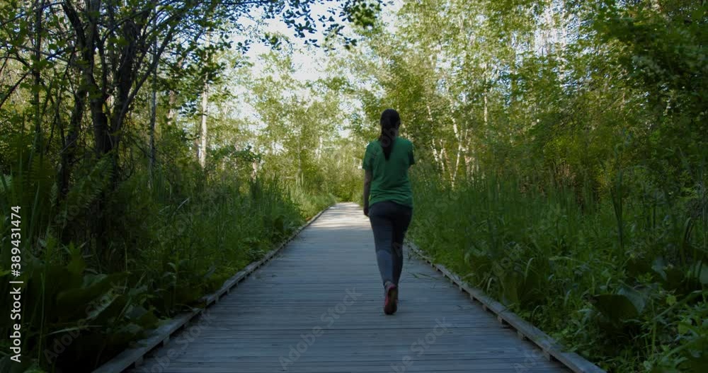 A girl walks down a wooden walkway amongst thick vegetation. As she walks into the distance, she enters bright sunlight. A girl walks down a wooden walkway amongst thick vegetation. As she walks into the distance, she enters bright sunlight.