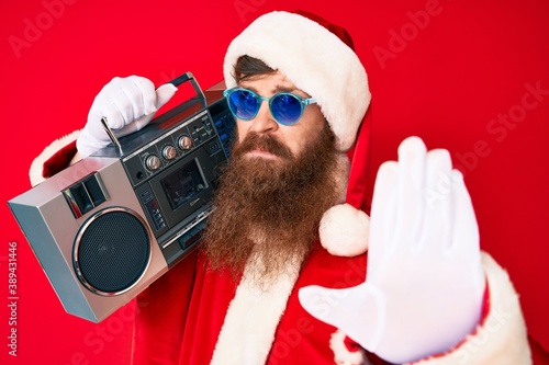 Handsome young red head man with long beard wearing santa claus costume and boombox with open hand doing stop sign with serious and confident expression, defense gesture