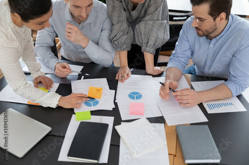 Top view focused young serious multiracial teammates involved in analyzing marketing research reports data statistics, developing company growth strategy or brainstorming ideas together at meeting.