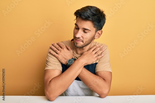 Young handsome man wearing casual clothes sitting on the table hugging oneself happy and positive, smiling confident. self love and self care © Krakenimages.com