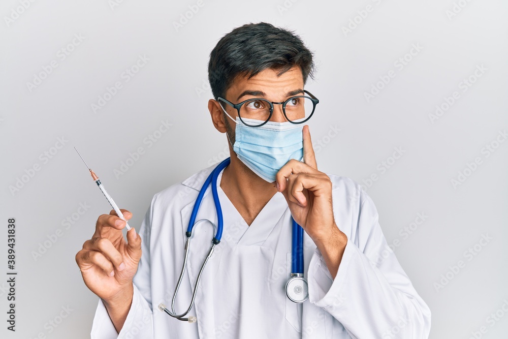 Young handsome man wearing doctor uniform and medical mask holding syringe serious face thinking about question with hand on chin, thoughtful about confusing idea Young handsome man wearing doctor uniform and medical mask holding syringe serious face thinking about question with hand on chin, thoughtful about confusing idea