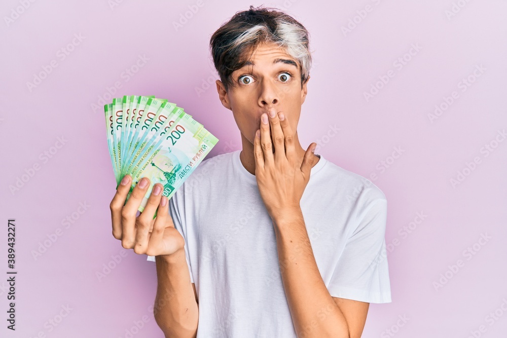 Young hispanic man holding russian 200 ruble banknotes covering mouth with hand, shocked and afraid for mistake. surprised expression Young hispanic man holding russian 200 ruble banknotes covering mouth with hand, shocked and afraid for mistake. surprised expression