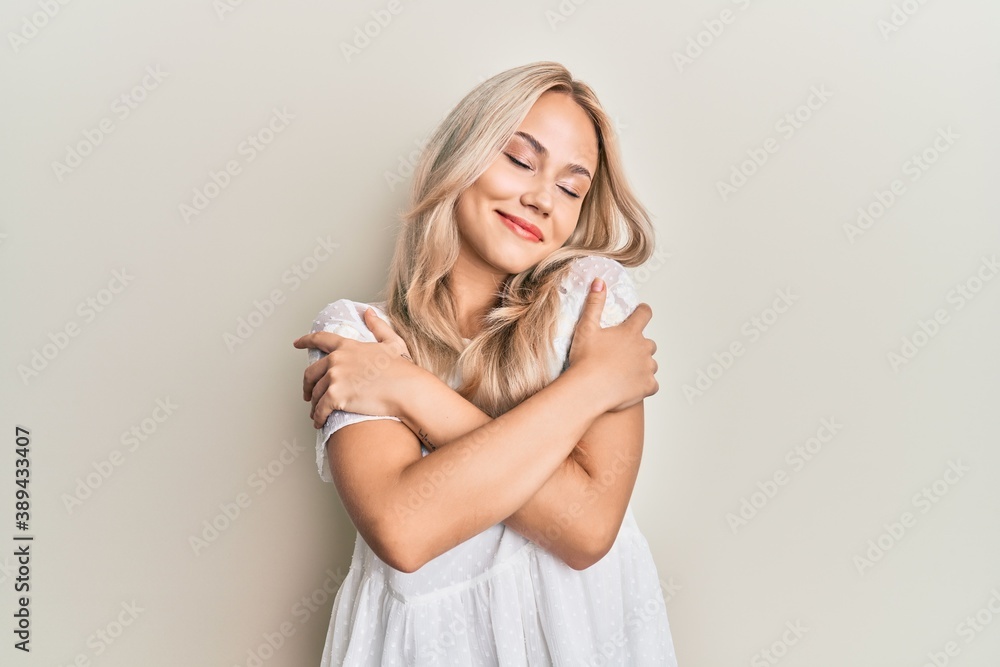 Beautiful caucasian blonde girl wearing white shirt hugging oneself happy and positive, smiling confident. self love and self care Beautiful caucasian blonde girl wearing white shirt hugging oneself happy and positive, smiling confident. self love and self care