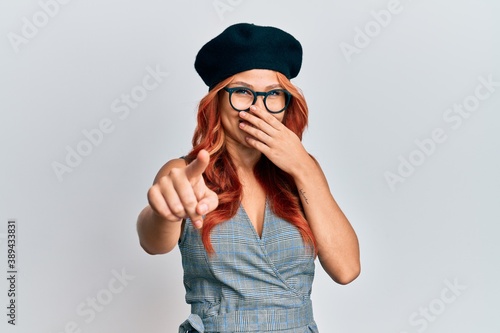 Young redhead woman wearing fashion french look with beret laughing at you, pointing finger to the camera with hand over mouth, shame expression