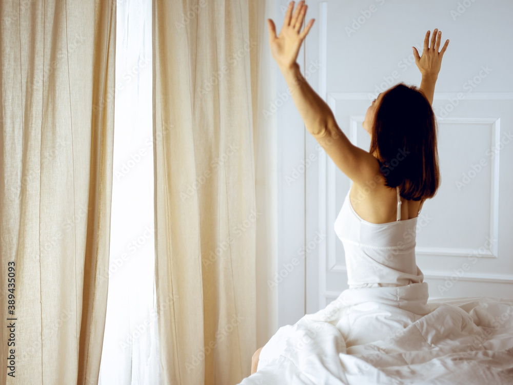 Fototapeta premium Woman stretching in bed after waking up, back view, entering a day happy and relaxed after good night sleep. Good morning and new day for brunette. Concept of Weekend, holidays