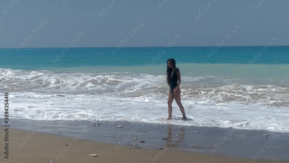 Petite Female in Swimsuit Walking on Rough Mediterranean Sea Waves Breaking on Beach, Full Frame Petite Female in Swimsuit Walking on Rough Mediterranean Sea Waves Breaking on Beach, Full Frame