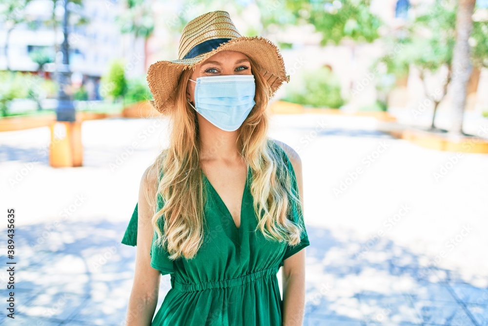 Young beautiful blonde woman on vacation wearing summer hat and medical mask. Standing with smile on face at street of city.