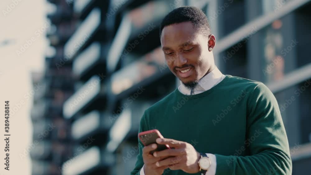 Confident african american man using mobile phone in modern city district. Positive business guy browsing financial news smiling standing outdoor. Successful adult.