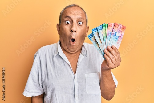 Handsome mature senior man holding swiss franc banknotes scared and amazed with open mouth for surprise, disbelief face photo