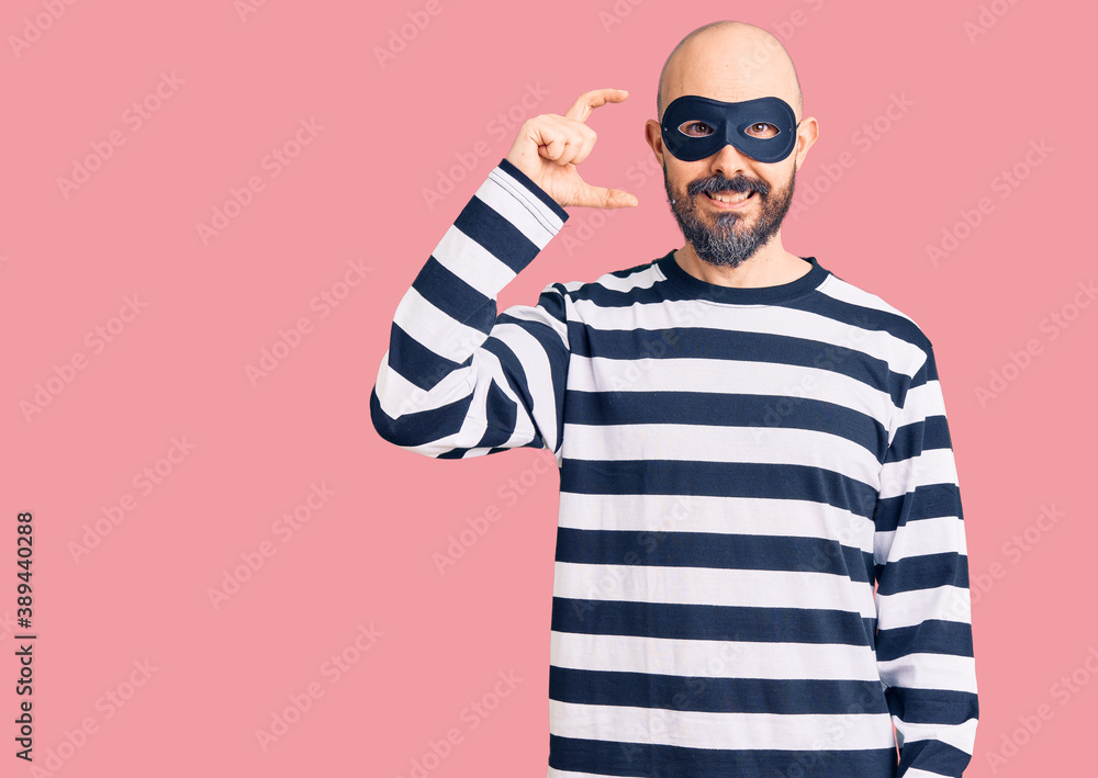 Young handsome man wearing burglar mask smiling and confident gesturing with hand doing small size sign with fingers looking and the camera. measure concept. Young handsome man wearing burglar mask smiling and confident gesturing with hand doing small size sign with fingers looking and the camera. measure concept.