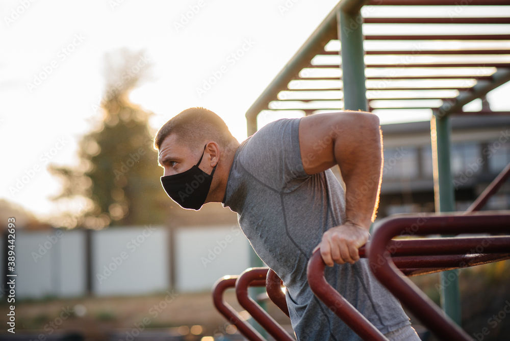 Fototapeta premium A young man does push-UPS, pull-UPS on a sports field in a mask during a pandemic at sunset. Sports, healthy lifestyle