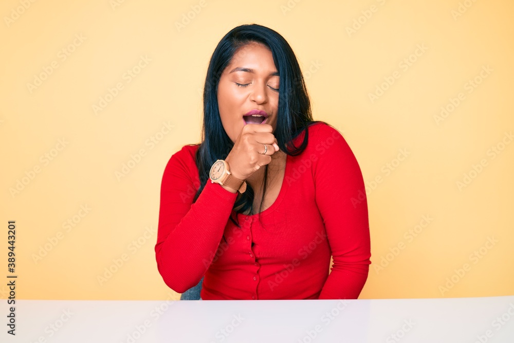 Beautiful latin young woman with long hair wearing casual clothes sitting on the table feeling unwell and coughing as symptom for cold or bronchitis. health care concept. Beautiful latin young woman with long hair wearing casual clothes sitting on the table feeling unwell and coughing as symptom for cold or bronchitis. health care concept.