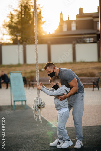 A father helps his son climb a rope on a sports field in masks during sunset. Healthy parenting and healthy lifestyle