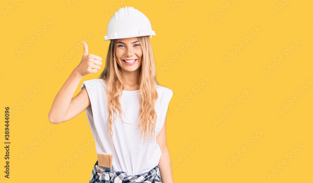 Beautiful caucasian woman with blonde hair wearing hardhat and painter clothes smiling happy and positive, thumb up doing excellent and approval sign