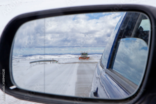 Snow plow cleaning a snow road seen from the rear view mirror of a car.