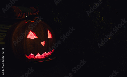 Side view of smiling Halloween pumpkin with eyes and mouth glowing inside on a house porch. Jack-o-lantern carved pumpkin for Halloween party. Dark background, copy space for text.