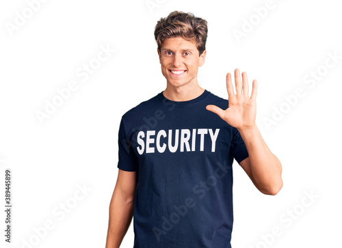 Young handsome man wearing security t shirt waiving saying hello happy and smiling, friendly welcome gesture