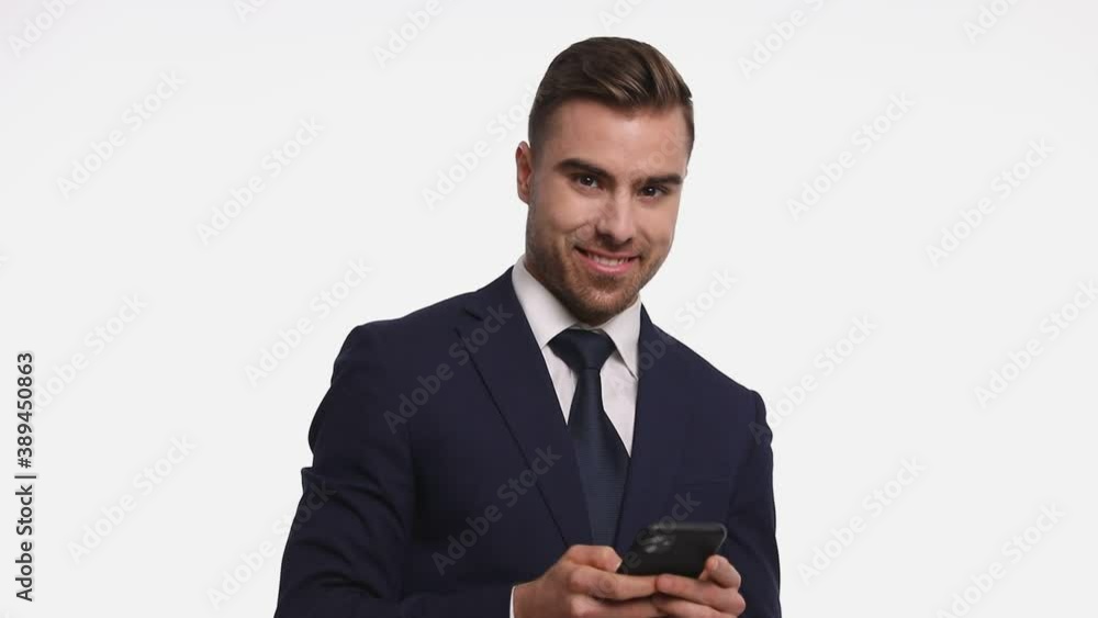 sexy young man wearing navy blue suit, holding phone, texting and smiling, making ok gesture, standing isolated on white background in studio sexy young man wearing navy blue suit, holding phone, texting and smiling, making ok gesture, standing isolated on white background in studio