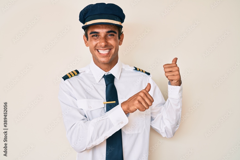 Young hispanic man wearing airplane pilot uniform pointing to the back behind with hand and thumbs up, smiling confident Young hispanic man wearing airplane pilot uniform pointing to the back behind with hand and thumbs up, smiling confident