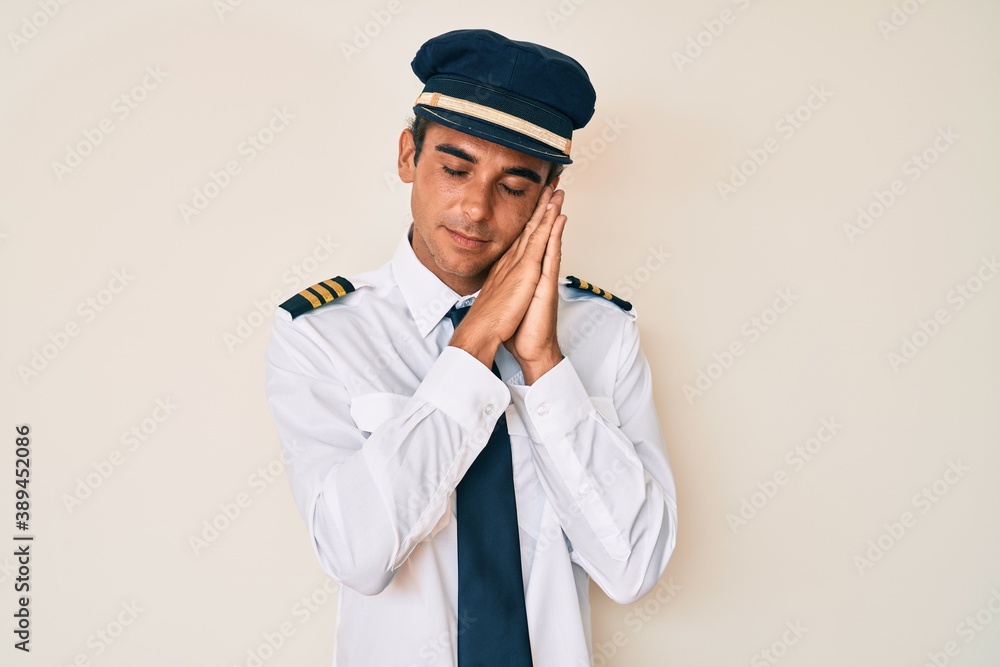 Young hispanic man wearing airplane pilot uniform sleeping tired dreaming and posing with hands together while smiling with closed eyes. Young hispanic man wearing airplane pilot uniform sleeping tired dreaming and posing with hands together while smiling with closed eyes.