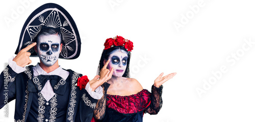 Young couple wearing mexican day of the dead costume over background confused and annoyed with open palm showing copy space and pointing finger to forehead. think about it.