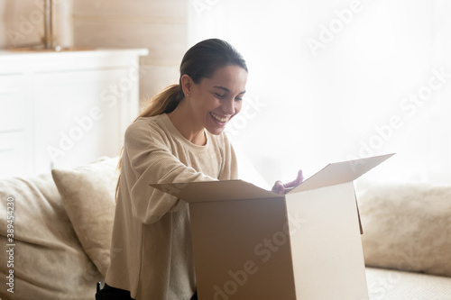 Let us look what is inside. Curious happy millennial woman client customer shopper buyer sitting on sofa at home opening unboxing cardboard box with mail package glad to get delivery easy and quick photo