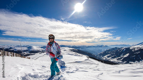A woman walking on powder snow with her snowboard on top of Katschberg in Austria. Panoramic view on the surrounding mountains. Winter wonderland. Sunny winter day. She is full of energy and happy