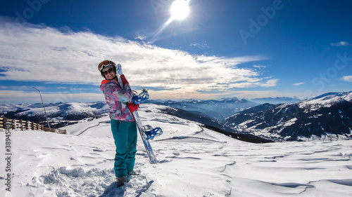 A woman walking on powder snow with her snowboard on top of Katschberg in Austria. Panoramic view on the surrounding mountains. Winter wonderland. Sunny winter day. She is full of energy and happy